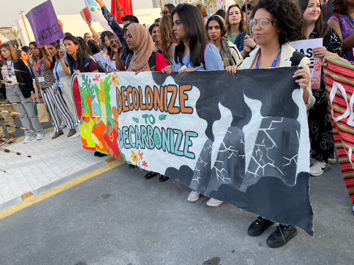 A group of students hold a colorful banner that reads 'decolonize to decarbonize' at COP. The title reads in white text 'Global Climate Talks and Tensions, Students and GW Alliance Director of Sustainability Research examine climate negotiations'